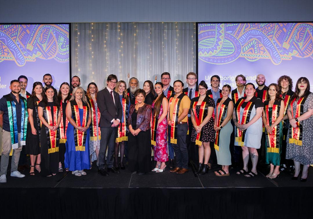 2023 Aboriginal and Torres Strait Islander Sashing Ceremony students pose on stage with Professor Kris Ryan, Deputy Vice-Chancellor (Academic), and Professor Bronwyn Fredericks, Deputy Vice-Chancellor (Indigenous Engagement).